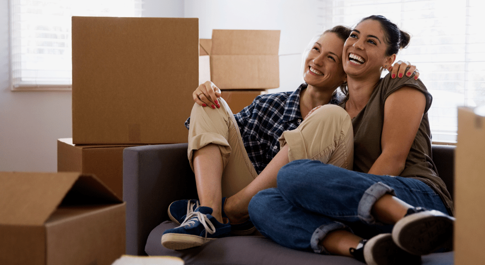 Couple smiling sitting on a couch together surrounded by moving boxes